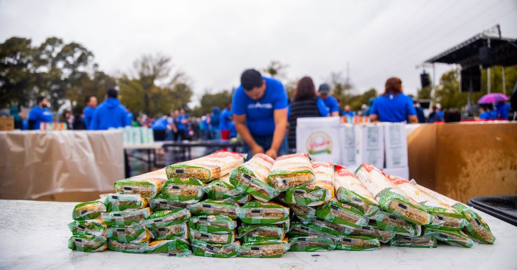 Stacked bags of rice on a table at an outdoor food drive with volunteers in blue shirts working behind